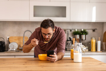 A man in a warm home kitchen enjoys a bowl of cereal while working on his laptop. He combines nutrition and productivity in a cozy, stylish setting, embracing a balanced daily routine.