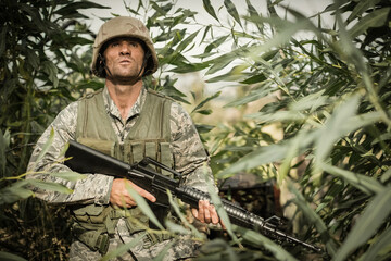 Mature male soldier patrolling jungle wearing camo uniform, helmet and vest, holding assault rifle