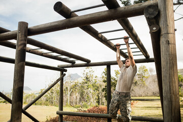 Male soldier with utility belt navigating monkey bars on grassy training ground under bright sky
