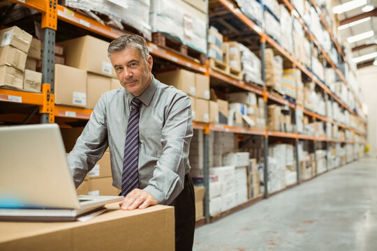 Mature adult man leaning on boxes and checking laptop in warehouse with metal shelving, copy space