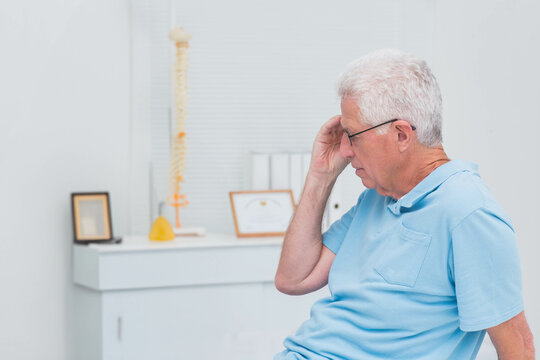 Senior man sitting in consultation room, wearing eyeglasses with spine model on cabinet, copy space
