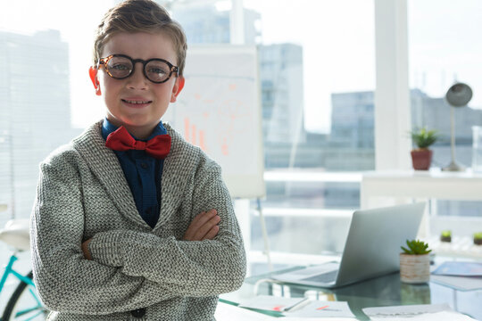 Boy standing with arms crossed in modern office beside open laptop, small plant and bicycle