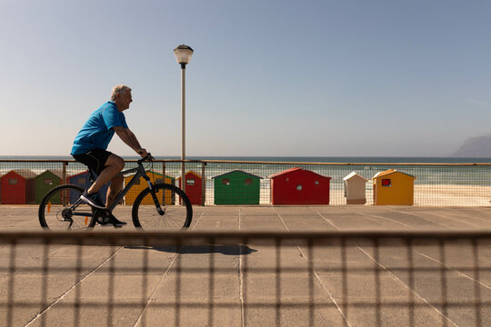 Senior man riding bicycle along seaside path past railing, beach huts and lamppost, copy space