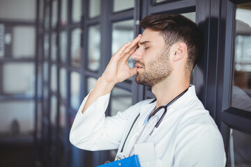 Male doctor leaning on glass wall in healthcare corridor, holding blue clipboard, pressing forehead