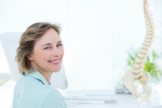 Female clinician showing spine model and typing on keyboard near plant in clinic, copy space