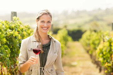 Woman standing in vineyard corridor wearing trench jacket holding red wine glass with sunlit vines