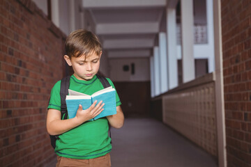 Boy student reading blue hardcover book in school corridor carrying black backpack near lockers