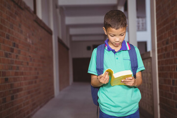 Boy standing in school corridor with brick walls reading yellow book and wearing blue backpack