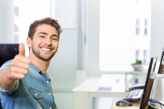 Man sitting at office desk wearing denim shirt giving thumbs up near computer setup, copy space