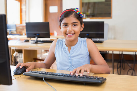 Indian schoolgirl wearing headband smiling at monitor in computer lab using keyboard and mouse