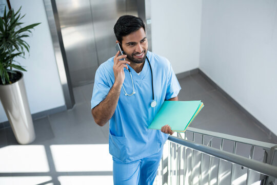 Asian man in scrubs wearing stethoscope climbing hospital stairwell holding smartphone and folders