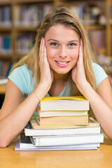 White teenager sitting at wooden table in library leaning on stack of textbooks by shelves smiling