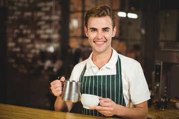 Smiling male barista pouring milk from pitcher into cup by espresso machine at coffee shop