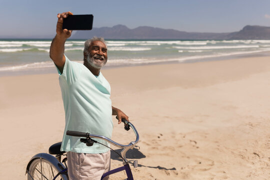 Senior Asian man standing on sandy beach beside bicycle holding smartphone taking selfie