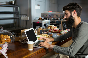Laptop sitting open beside steaming paper coffee cup on wooden counter under glass pastry domes