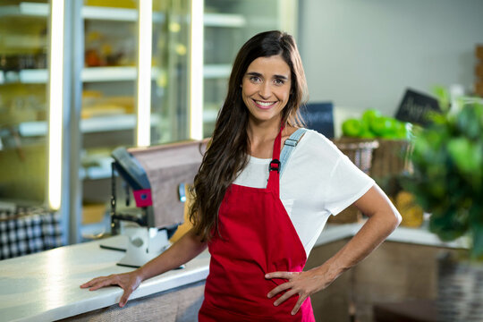 Female store clerk leaning on service counter wearing red apron in grocery shop near digital scale