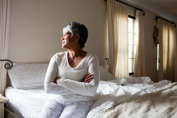 Senior woman sitting on unmade bed in bedroom crossing arms and gazing through curtains, copy space © WavebreakMediaMicro