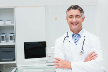 Male doctor wearing lab coat, stethoscope crossing arms in office with computer monitor, copy space