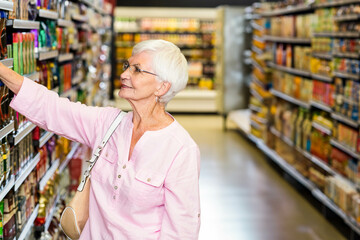 Senior woman reaching for boxed goods on supermarket aisle shelves while wearing glasses and purse