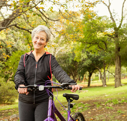 Senior woman standing under leafy tree in park holding purple bicycle handlebars sporting jacket