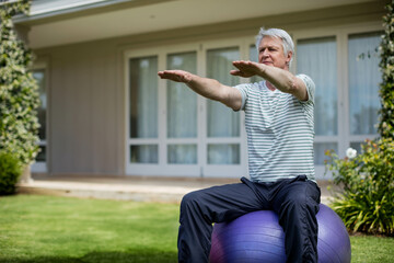 Senior male balancing on purple stability ball on lawn outside home in workout clothes, copy space