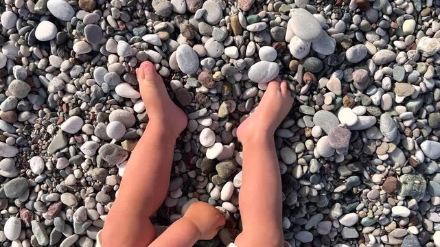 Baby feet on the beach among stones. A toddler plays sitting on pebbles by the sea. Vacation with family.