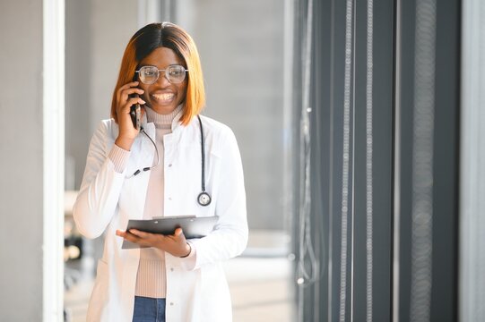 African american female doctor wearing lab coat talking on smartphone in hospital corridor - Powered by Adobe