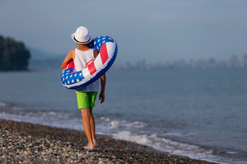 A boy walks along the beach with an inflatable ring in the colors of the American flag on a summer...