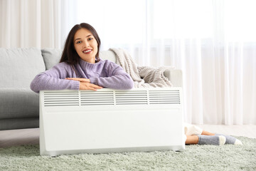 Obraz premium Young woman relaxing at home with electric heater, cozy indoor comfort and warmth during cold weather, the heating appliance provides a comfortable environment in the living room