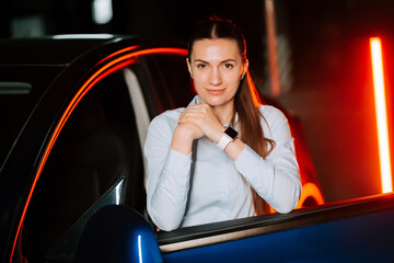 Young woman poses confidently by her car at night in a modern urban setting