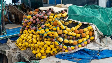 Weathered yellow and brown fishing floats and tangled nets piled on a dockside boat, with parts of the vessel and harbor scene in the background. © CanYalicn