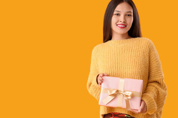 Beautiful young Asian woman with gift box on yellow background