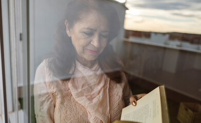 Senior woman reading a book by the window at sunset