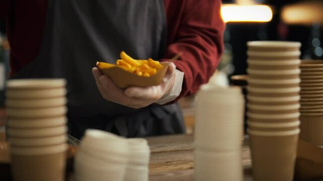 An experienced cafe worker quickly and professionally puts ready-made fresh French fries into environmentally friendly cardboard dishes. The atmosphere of a cozy fast food cafe. Visitors order food