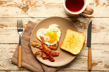 Plate with tasty bacon, cup of tea, toast, fried egg and cutlery on light wooden background