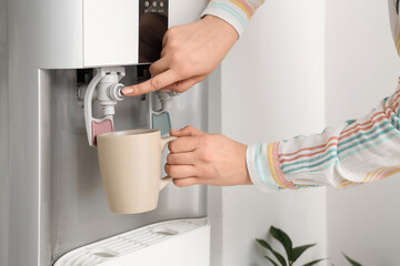 Woman filling cup with filtered water from cooler near white wall
