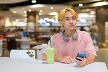 Thai gay teen man with headphones using phone and drinking iced latte green tea in modern cafe