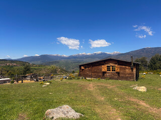 A wooden cabin with Snow-Capped Peaks in the mountains on a sunny day. Rustic Cabin. Scenic view of rolling hills, mountains, and a cloudy sky under sunlight.