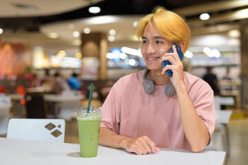 Thai gay teen man with headphones using phone and drinking iced latte green tea in modern cafe