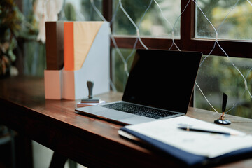 Modern minimalist workspace with laptop, document folders, and rubber stamp on wooden table beside a large window. Bright, serene home office setup.