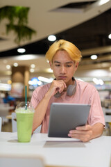 Thai gay teen man with headphones using digital tablet and drinking iced latte green tea
