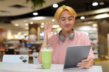 Thai gay teen man with headphones using digital tablet and drinking iced latte green tea