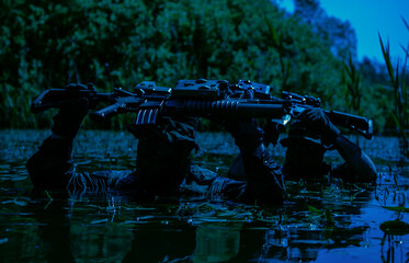 Two armed soldiers in camouflage, carrying rifles above water, wade slowly through a dense, swamp-like area surrounded by greenery.
