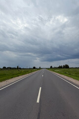 Asphalt road in the countryside running through green fields on a cloudy day. Rural landscape with road and storm clouds