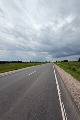 Asphalt road in the countryside running through green fields on a cloudy day. Rural landscape with road and storm clouds