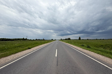 Asphalt road in the countryside running through green fields on a cloudy day. Rural landscape with road and storm clouds
