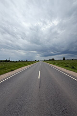Asphalt road in the countryside running through green fields on a cloudy day. Rural landscape with road and storm clouds