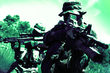 Two soldiers in camouflage uniforms and helmets aiming rifles in a wooded outdoor setting, under a bright sky.