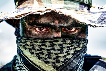 Close-up of a person with camouflage face paint and headscarf, wearing a patterned hat and looking directly at the camera with intense eyes.