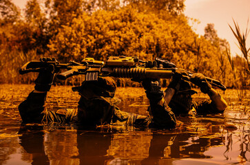 Two camouflaged soldiers with rifles raised above water, navigating through a swampy area with dense foliage in the background.